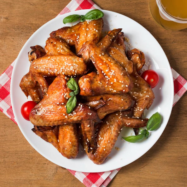A white plate of sesame chicken wings with basil and cherry tomatoes on a wooden table.