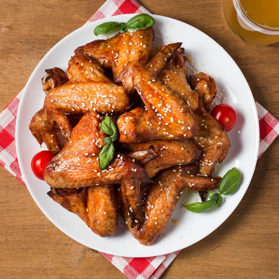 A white plate of sesame chicken wings with basil and cherry tomatoes on a wooden table.