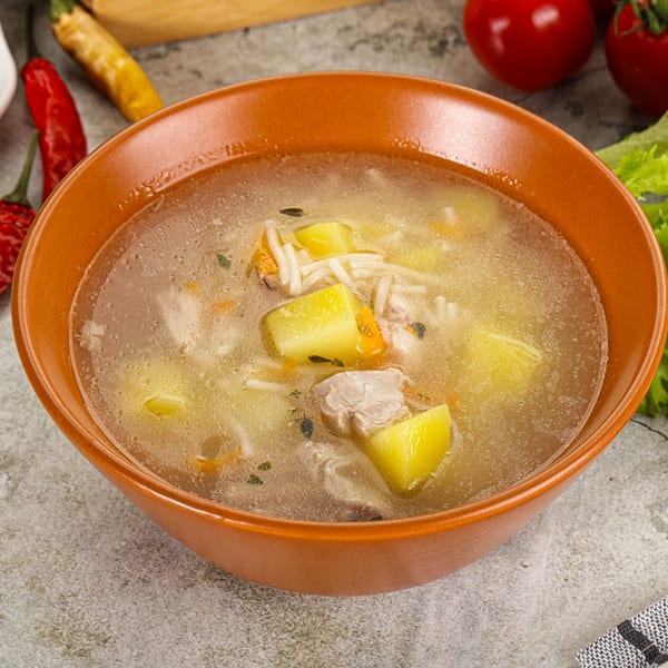 A bowl of clear chicken noodle soup with potatoes and meat, garnished with red chilies.